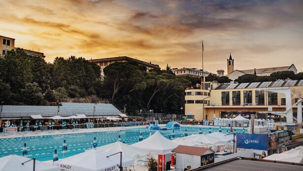 Area piscina con lettini e ombrelloni, circondata da edifici e alberi.