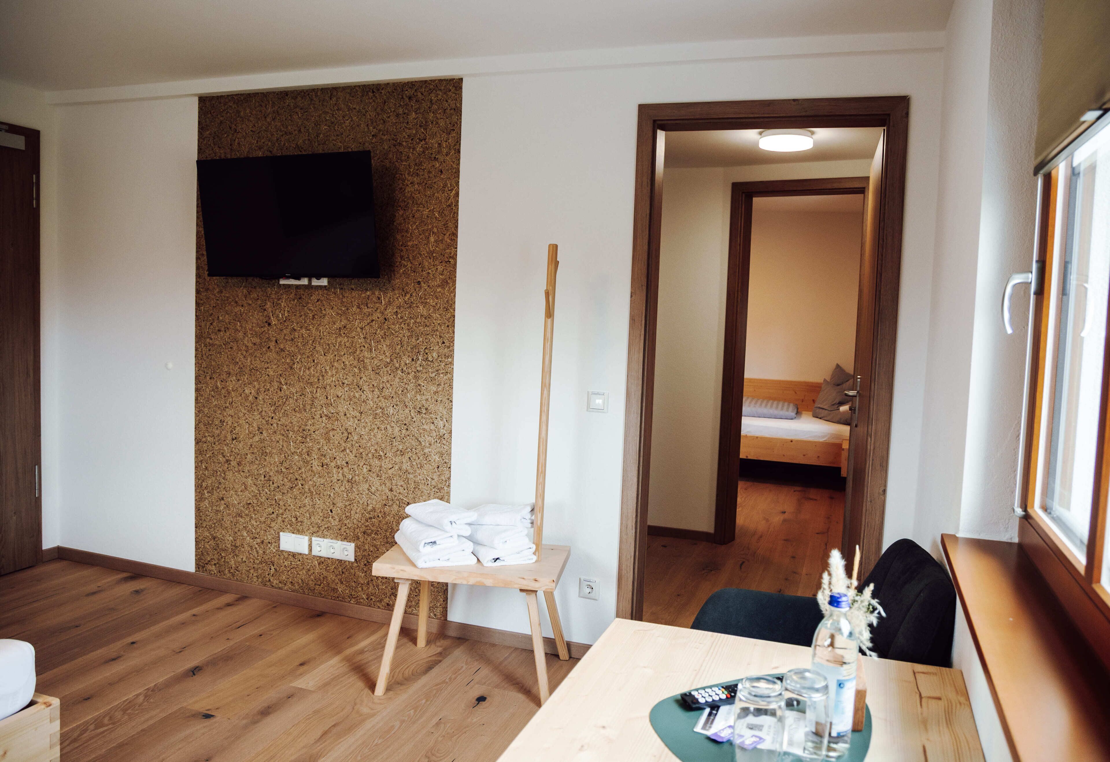Een hotelkamer met een houten vloer, een televisie bevestigd aan een kurkwand, een klein houten tafeltje met handdoeken, en een bed met een houten hoofdeinde.