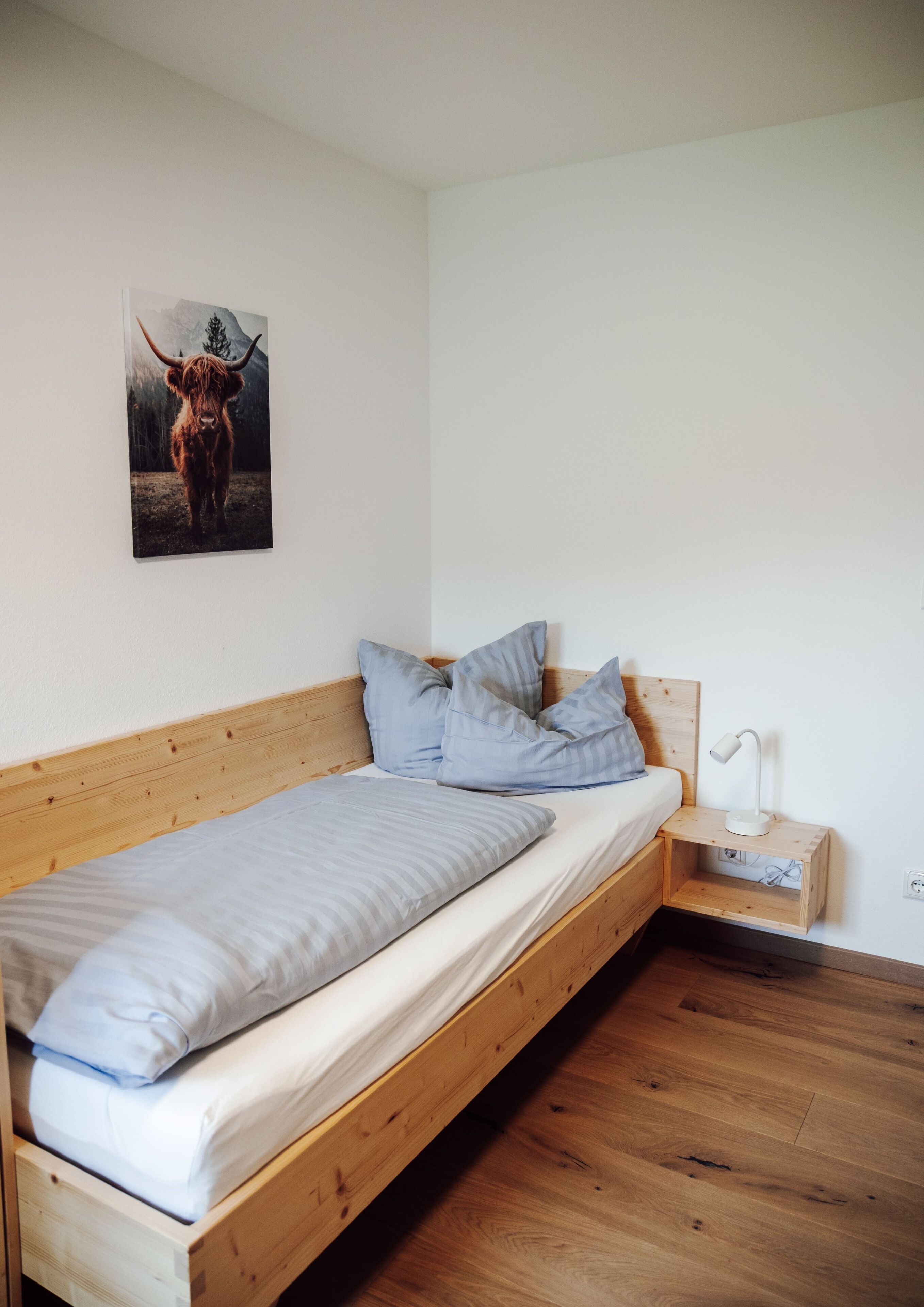 A single bed with a wooden frame, a white mattress, and two pillows. There is a bedside table with a lamp and a power outlet. A framed picture of a yak is on the wall.