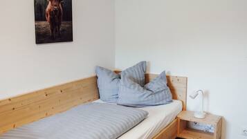 A single bed with a wooden frame, a white mattress, and two pillows. There is a bedside table with a lamp and a power outlet. A framed picture of a yak is on the wall.