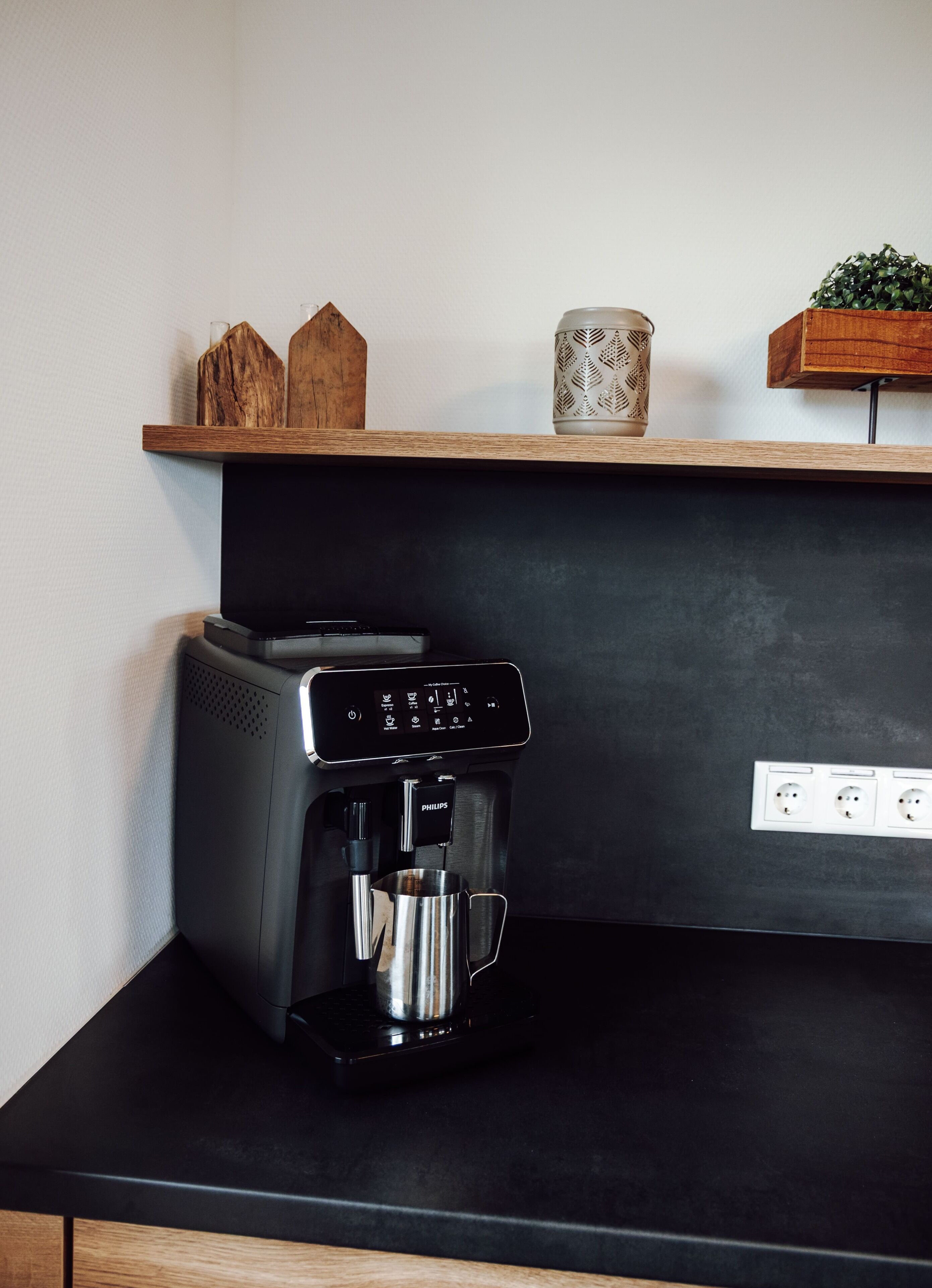 A coffee machine on a black countertop with a wooden shelf above it.