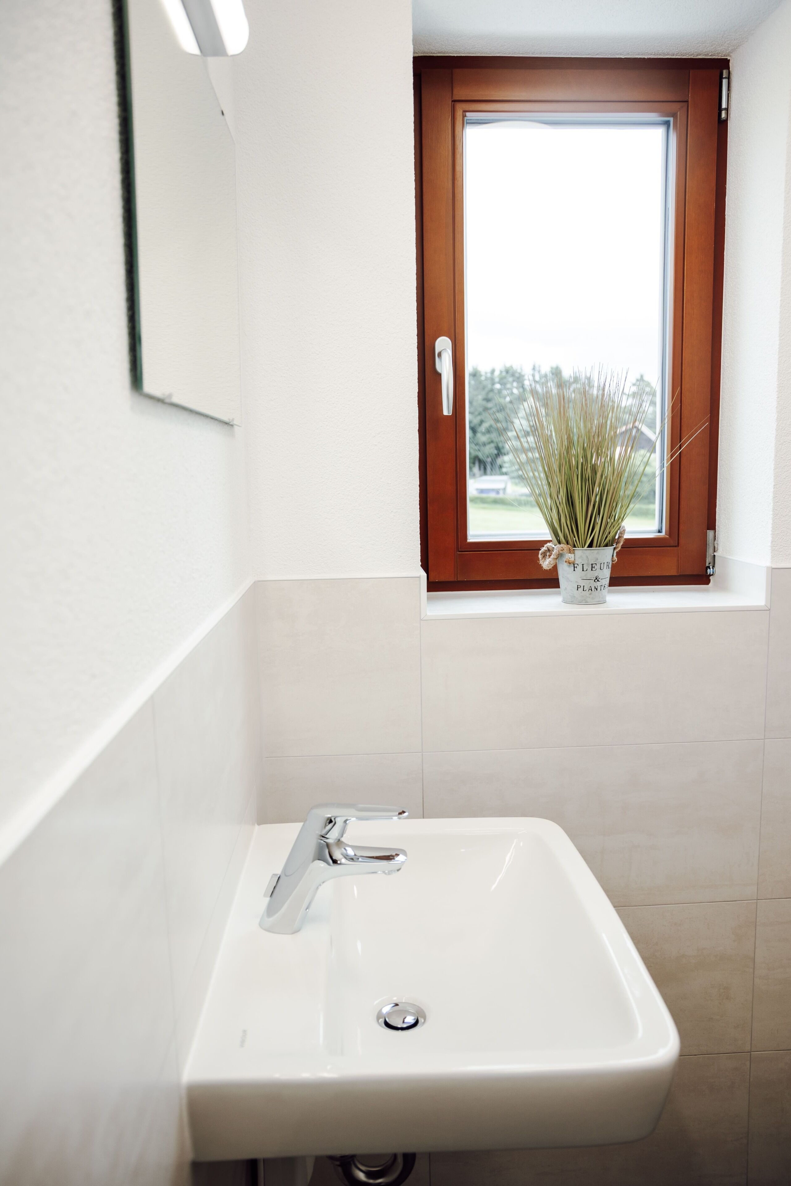 A bathroom with a white sink, a window with a wooden frame, and a potted plant on the windowsill.