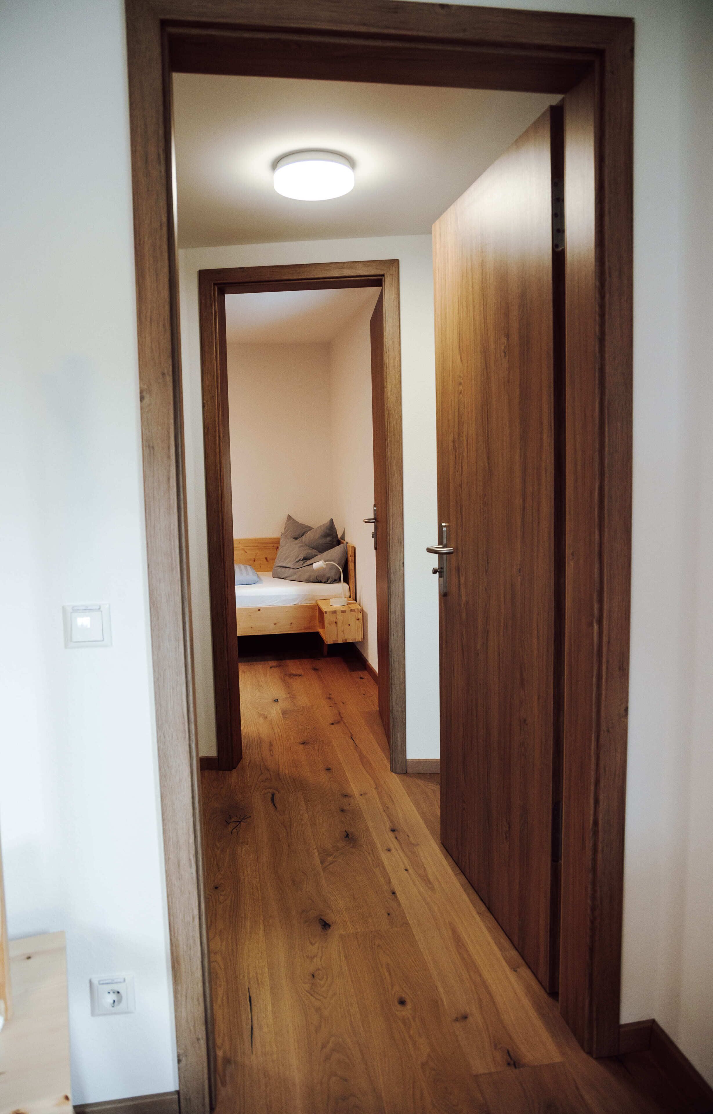 Un couloir étroit mène à une chambre dotée d’un plancher en bois, d’un lit et d’une table de chevet.