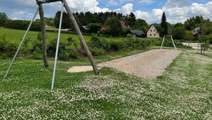 A playground with swings and a gravel area, surrounded by green grass and white flowers, with houses and trees in the background. - Das Fränkische Reichenbach (Reichenbach)