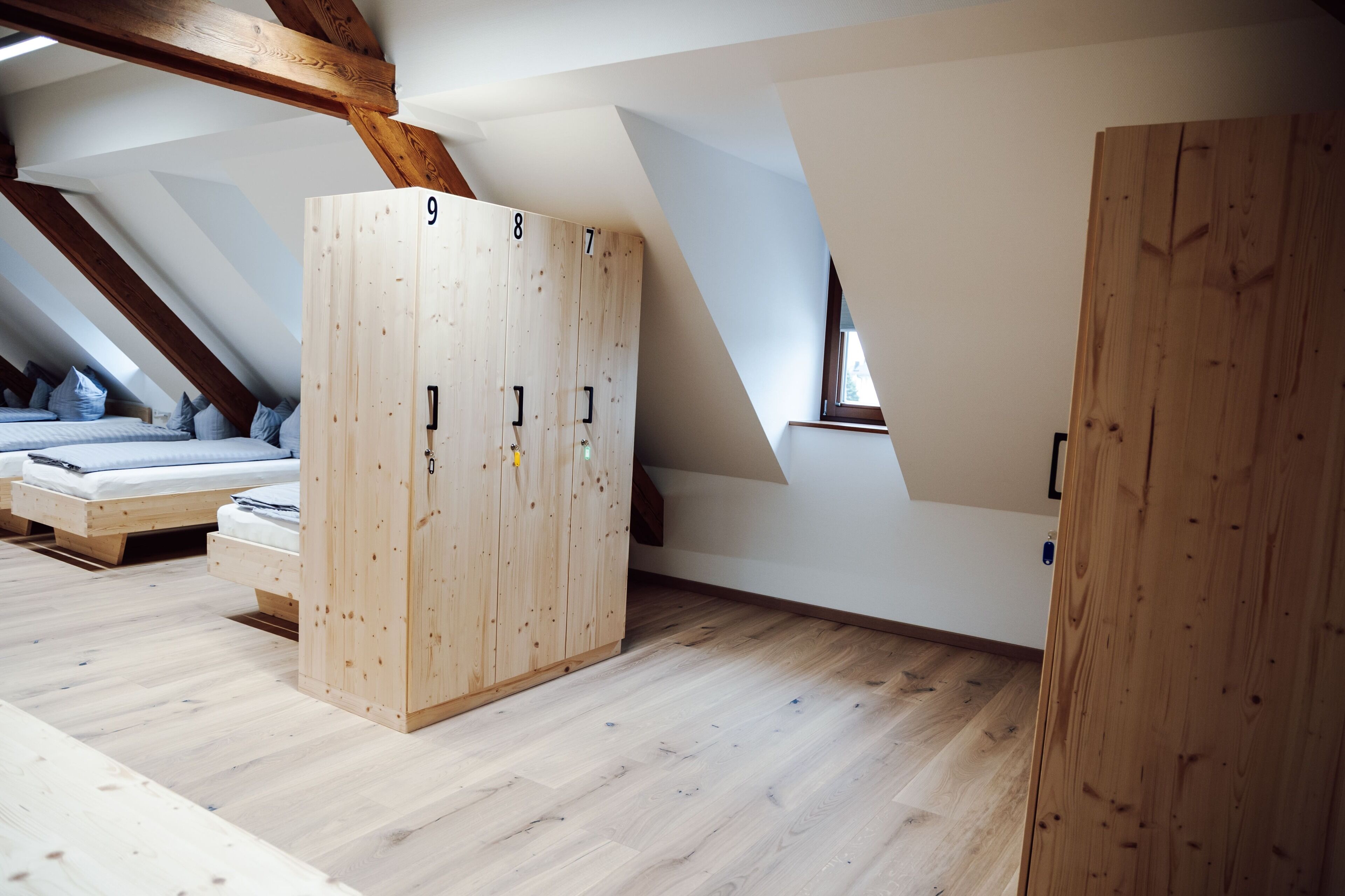 Ein Schlafzimmer mit schrägem Dach, Holzboden, einem großen Kleiderschrank mit vielen Fächern und einem Bett mit blauer Bettwäsche.