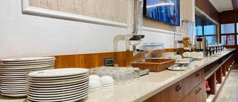 Una barra de buffet con platos apilados, vasos y dispensador de bebidas.
