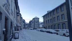 Una calle nevada flanqueada por edificios, coches aparcados y una montaña al fondo.