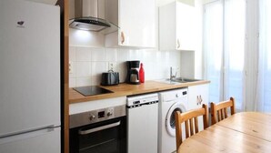 Cocina compacta equipada con refrigerador, horno, lavavajillas y lavadora. Cuenta con una mesa de comedor de madera y sillas.