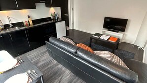 Una sala de estar moderna con un sofá de cuero negro, una mesa de comedor con sillas blancas, una zona de cocina con armarios negros y un televisor de pantalla plana sobre un pedestal.