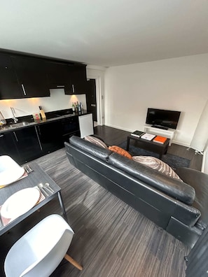 Ein modernes Wohnzimmer mit einer schwarzen Ledersofa, einem Esstisch mit weißen Stühlen, einer Küchenzeile mit schwarzen Schränken und einem Flachbildfernseher auf einem Standfuß.