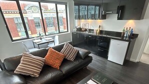 Una sala moderna con un sofá de cuero negro, una mesita de centro con libros y vistas a una cocina y a un paisaje urbano a través de ventanas amplias.
