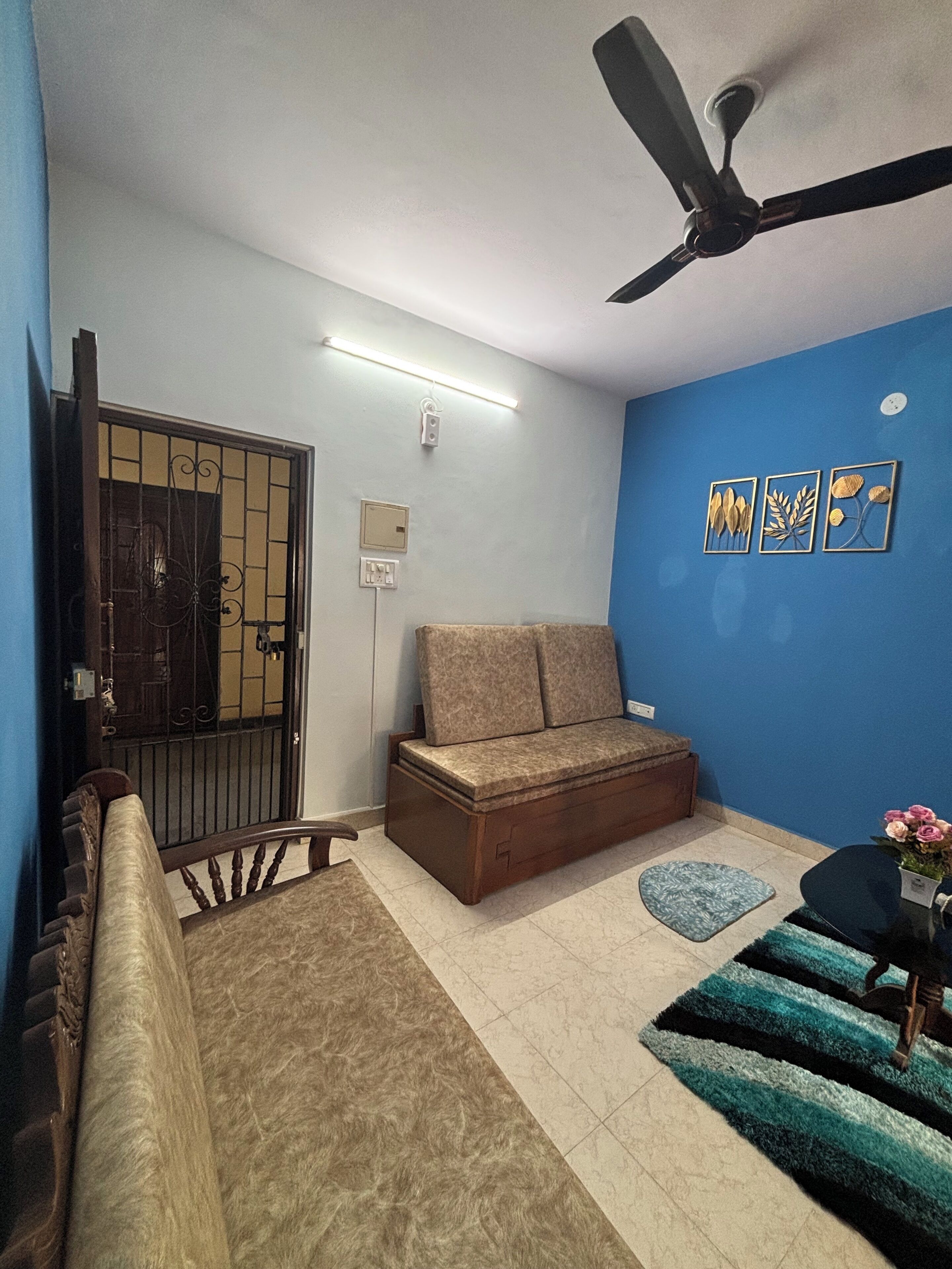 Una habitación con sofá, ventilador de techo, luz empotrada, una puerta y una pequeña mesa con un florero.