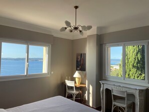 Ein Schlafzimmer mit einem Bett, einem Schreibtisch mit Stuhl, einer Lampe, einem Kamin und großen Fenstern mit Blick auf das Meer und viel Grün.