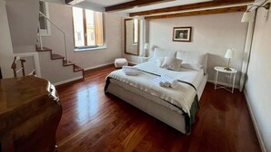 A bedroom with a wooden floor, a bed with white linens, a nightstand with a lamp, a window with a view, and a staircase. - Penthouse23 Vatican Charming Apartment (Roma)