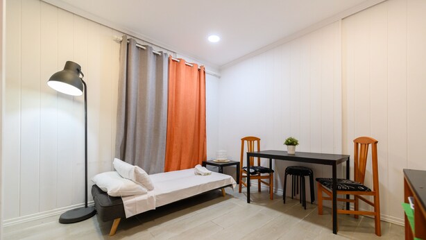 Una habitación pequeña y bien iluminada con una cama individual, un escritorio con dos sillas, una mesita con una planta y una lámpara de pie.
