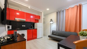Cocina compacta con armarios rojos, lavadora y microondas. Hay un sofá gris y una mesita con una lámpara.