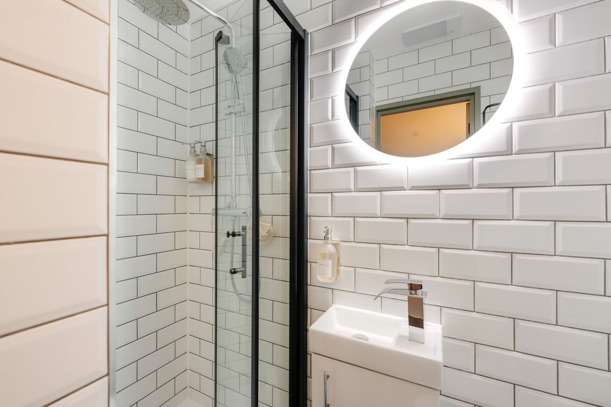 Un baño con azulejos blancos tipo metro, ducha con cerramiento de vidrio, espejo redondo y lavamanos blanco.