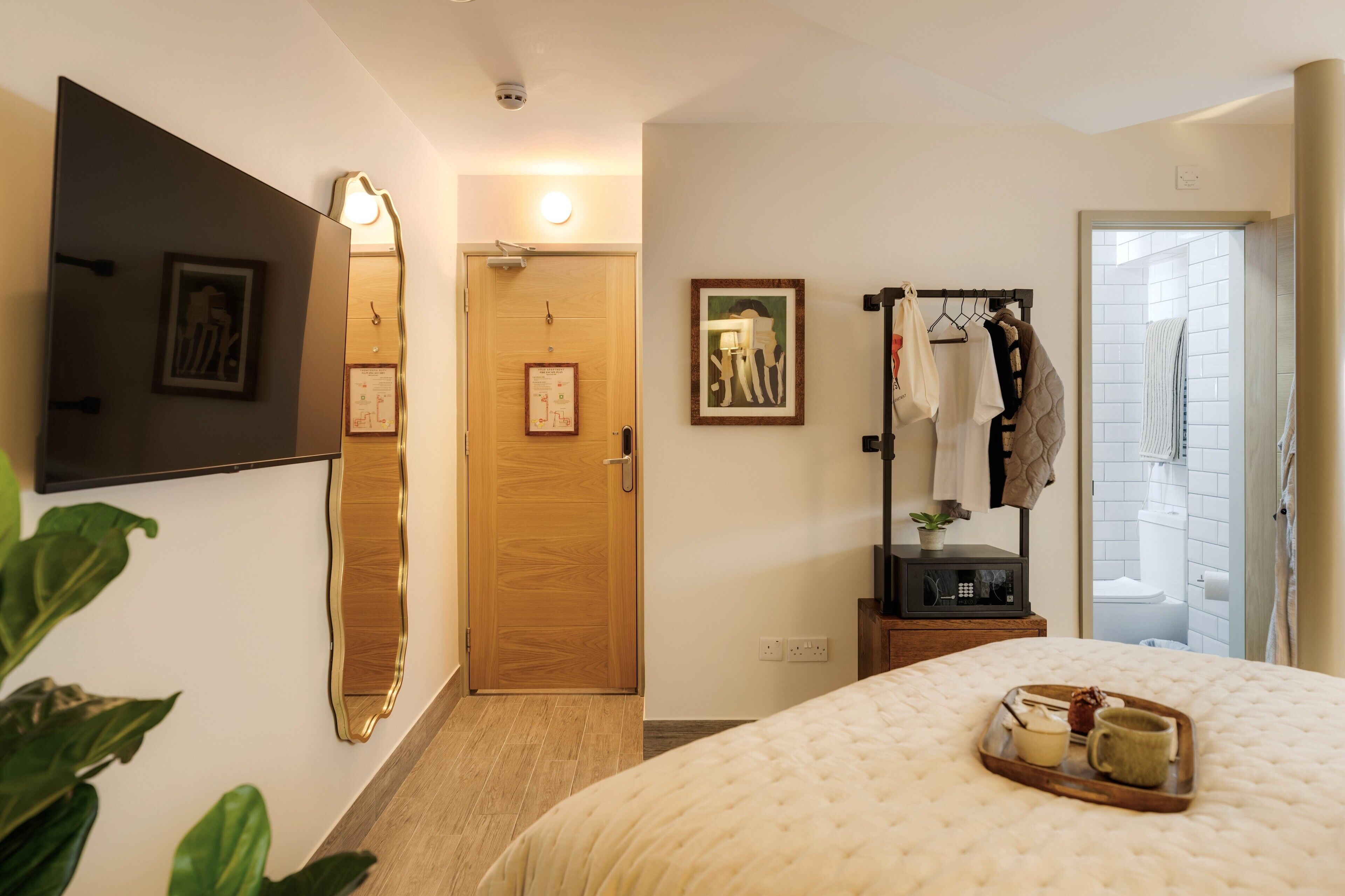 Een hotelkamer met een bed, een nachtkastje met een dienblad, een spiegel, een kapstok en een deur naar de badkamer.