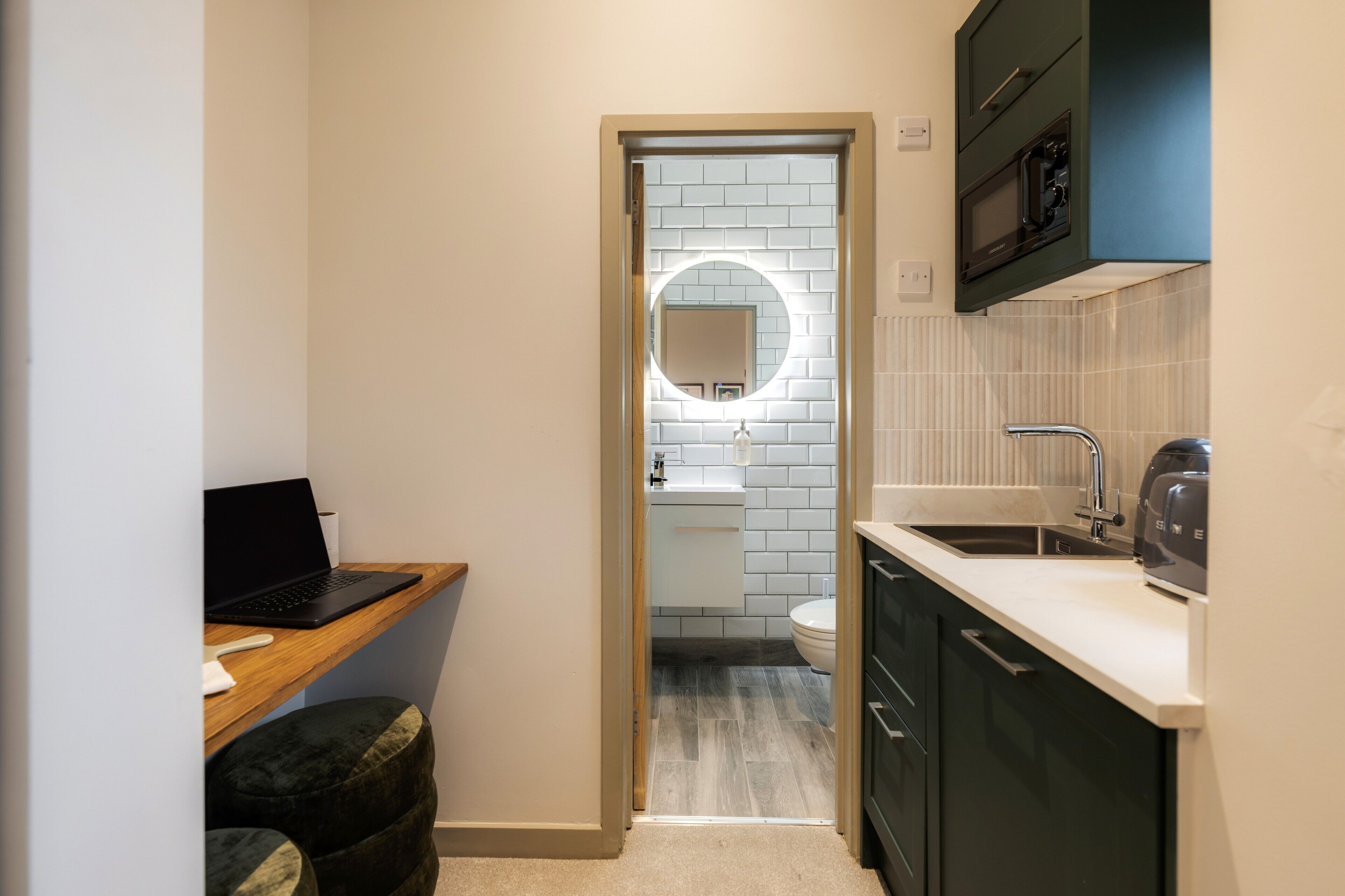 A compact kitchen with a microwave, sink, and a laptop on a desk.
