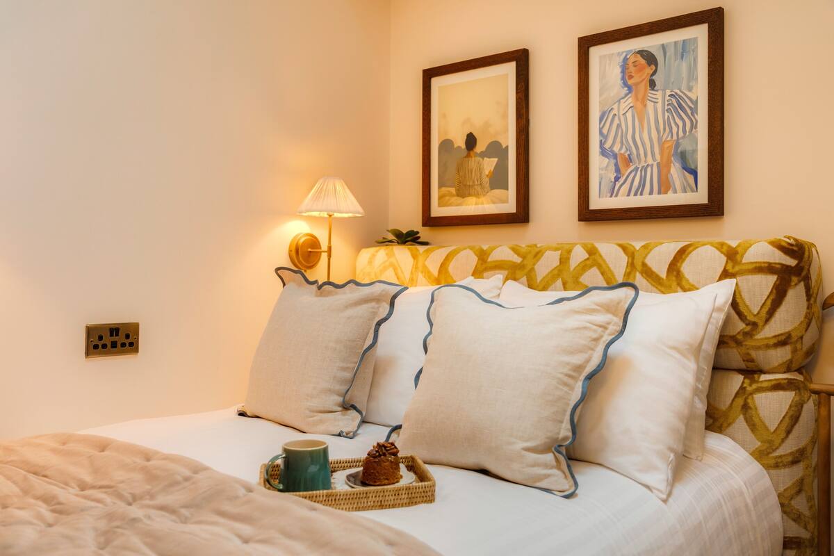 A neatly made bed with a tray of pastries and a mug, a wall-mounted lamp, and two framed artworks above the bed.