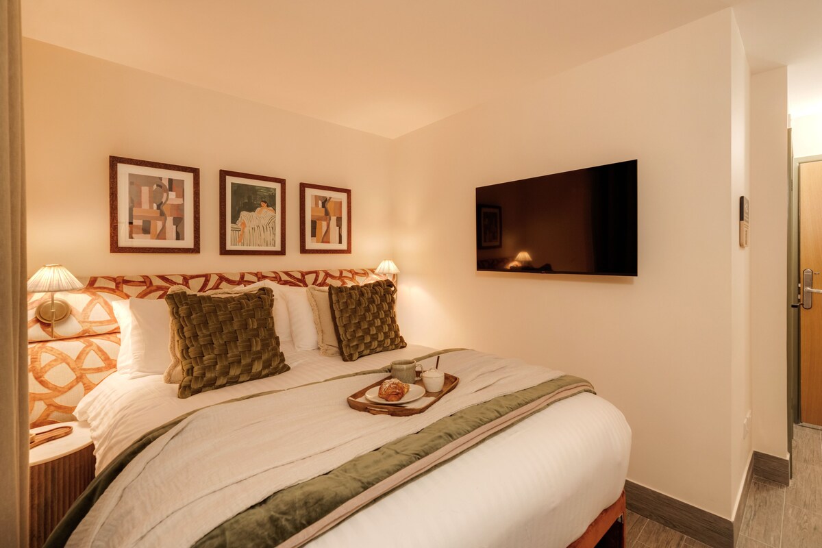 A neatly made bed with a tray of breakfast items, a flat-screen TV mounted on the wall, and three framed artworks above the bed.