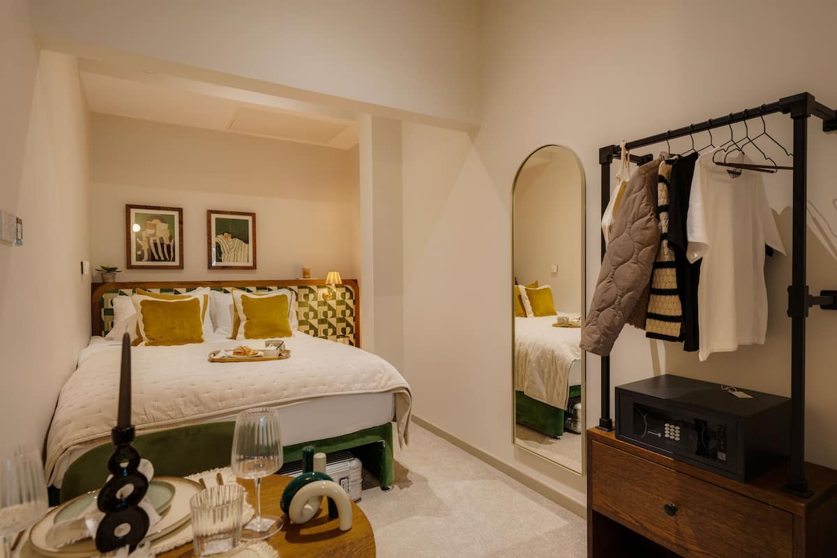 A hotel room with a bed, bedside table, mirror, and a clothing rack.