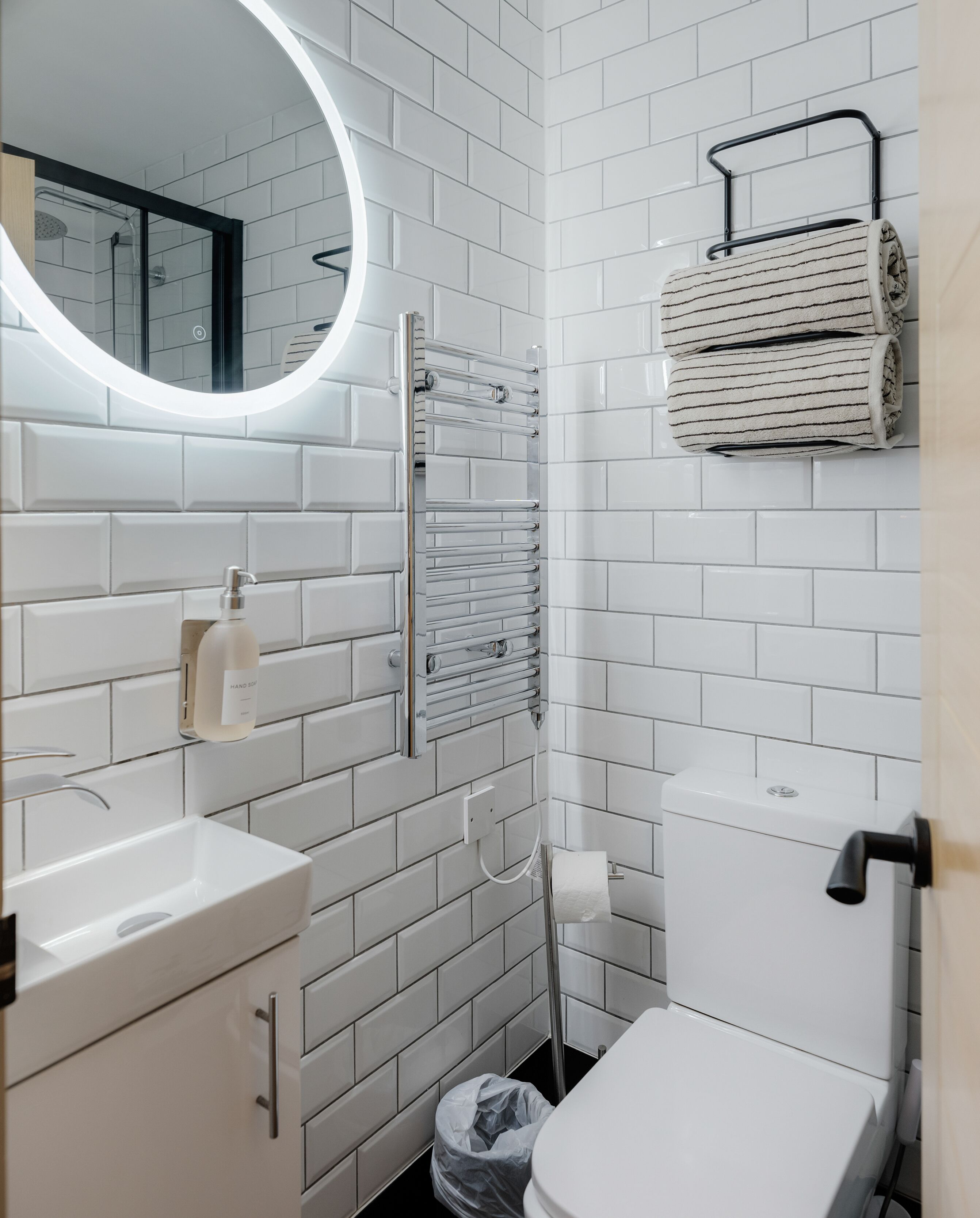 Un baño moderno con azulejos blancos tipo metro, un espejo redondo con luz, un perchero, un inodoro y un dispensador de jabón para manos.