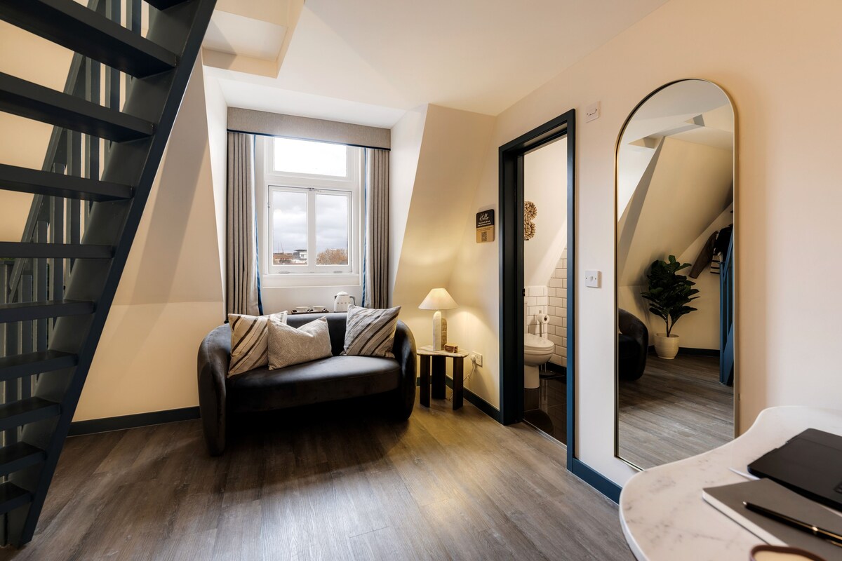 A modern living room with a dark leather sofa, a small side table with a lamp, a large mirror, and a staircase.