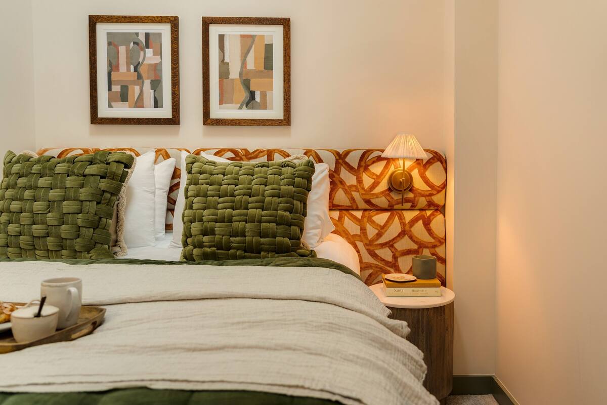 A neatly made bed with green and white pillows, a bedside table with a lamp and a book, and two framed abstract paintings on the wall.