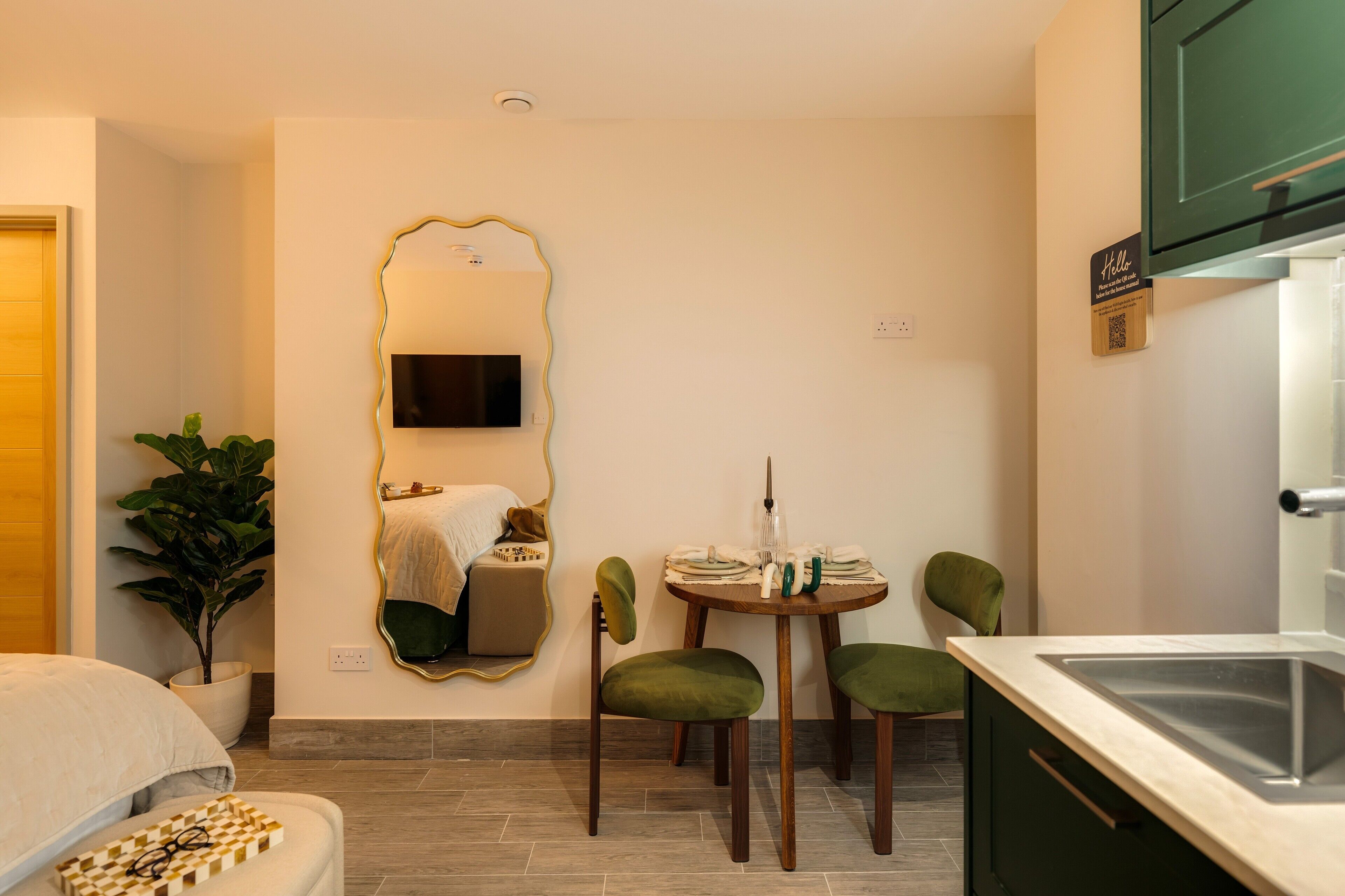 A compact room with a bed, a dining table with chairs, a mirror, a TV, and a sink.