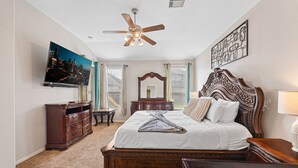 A bedroom with a bed, a ceiling fan, a TV, a dresser, a mirror, and a window with curtains.