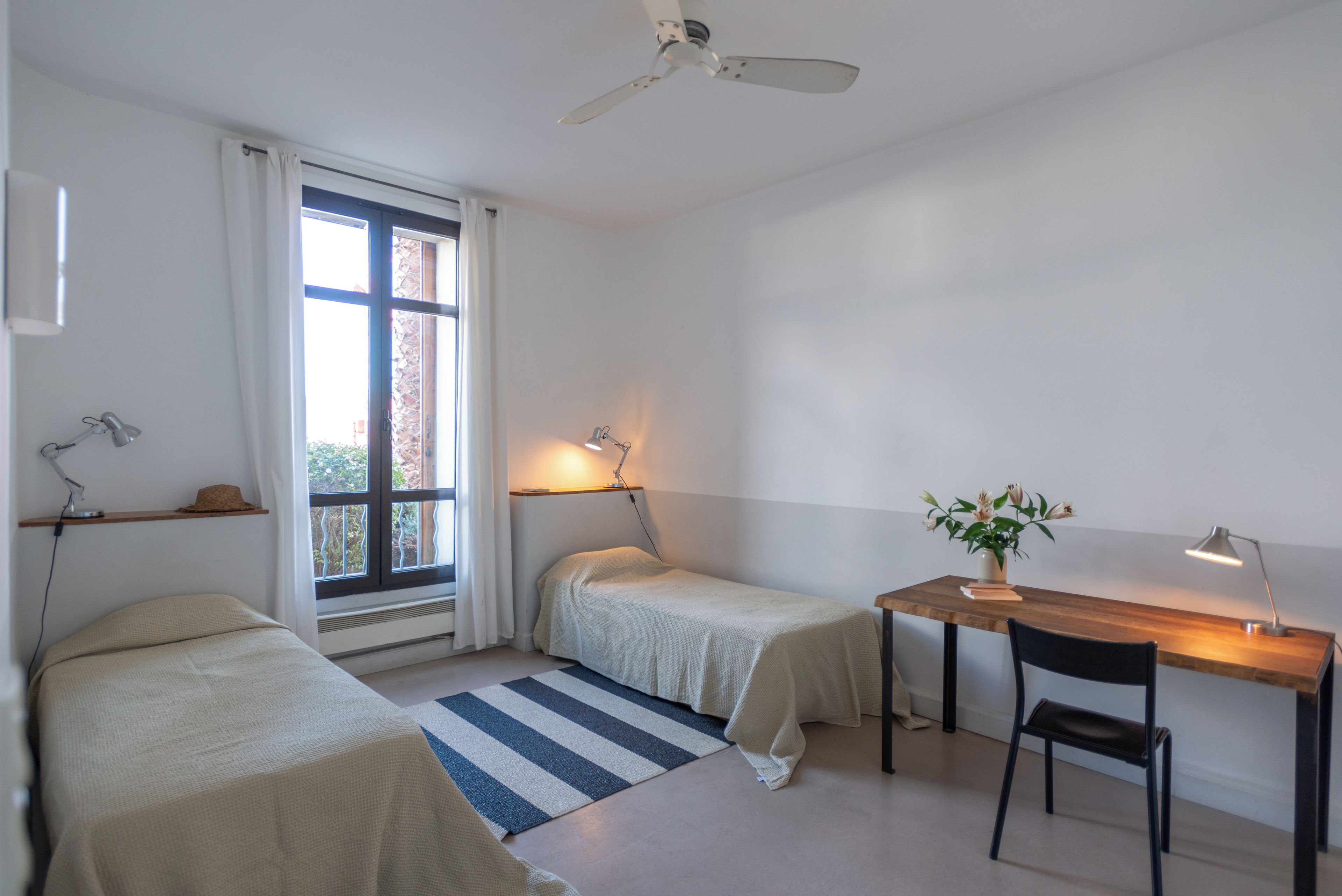 Une chambre avec deux lits, un bureau, une chaise, un ventilateur de plafond et une fenêtre avec des rideaux.