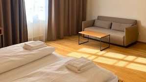 Quarto de hotel com cama, sofá, mesa de centro e uma grande janela com cortinas.