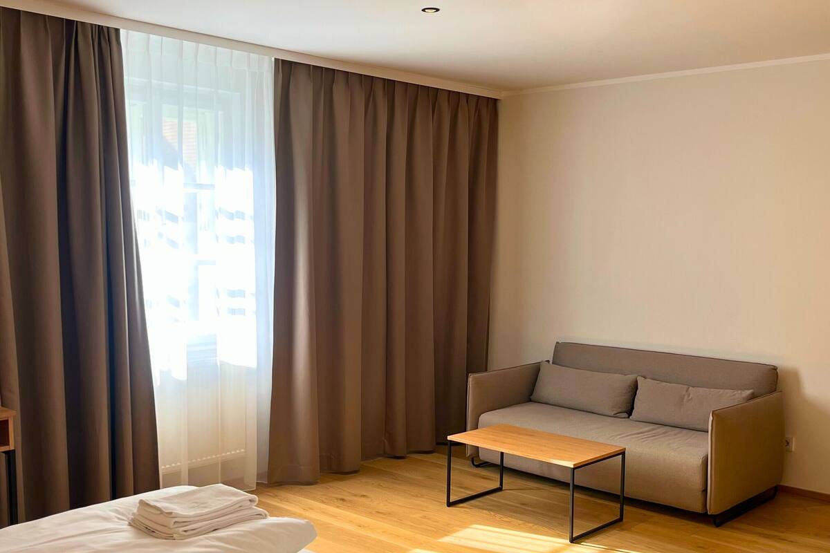 Camera d'albergo con un letto, una poltrona letto, un tavolino da caffè e un'ampia finestra con tende.