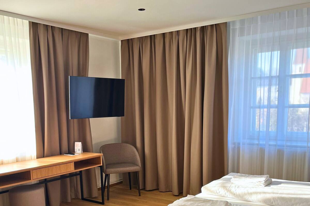 Una camera d'albergo con pavimento in legno, un letto con lenzuola bianche, una scrivania con una sedia, una televisione e tende.