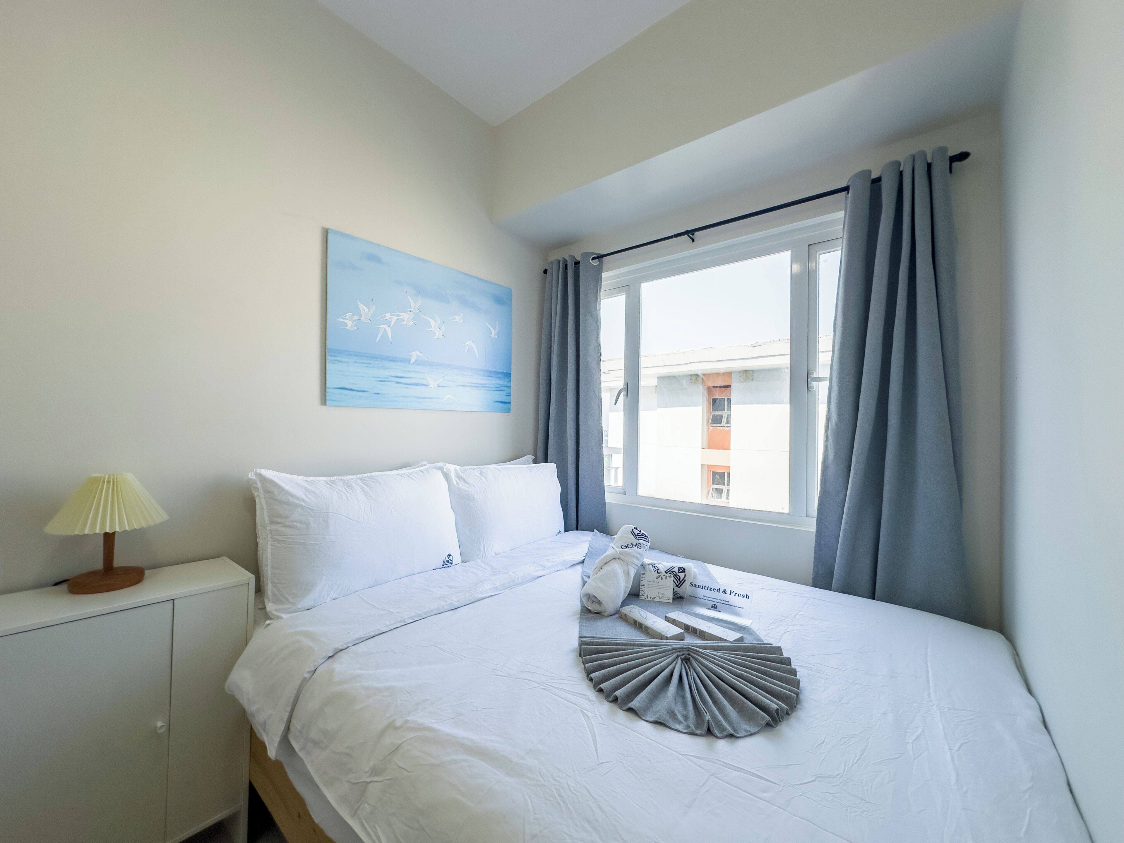 A bedroom with a bed, a bedside table with a lamp, a window with curtains, and a painting on the wall.