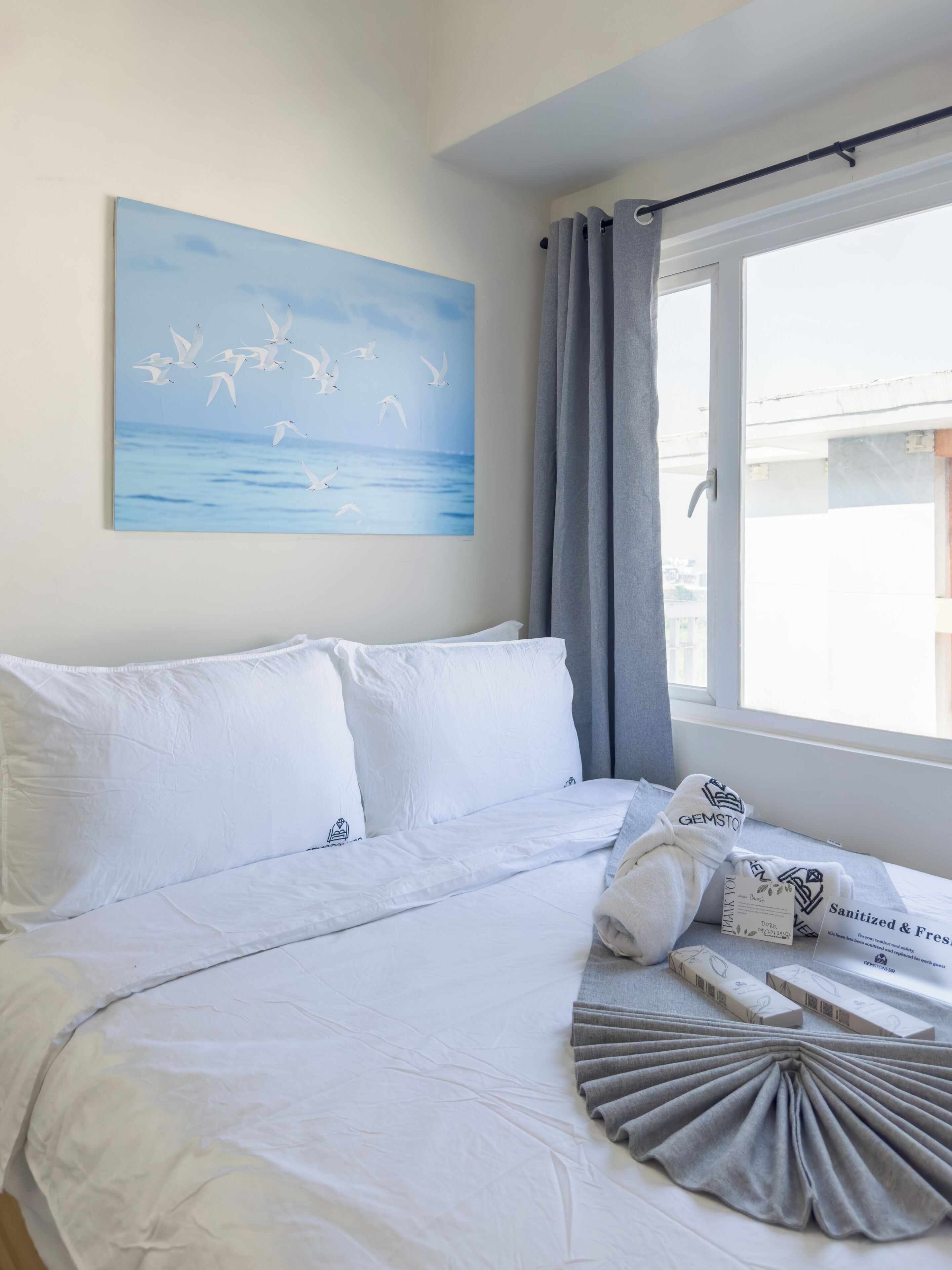 Ein Schlafzimmer mit einem Bett, Kissen, einer Decke, einem Fenster mit Vorhängen und einem Bild an der Wand.