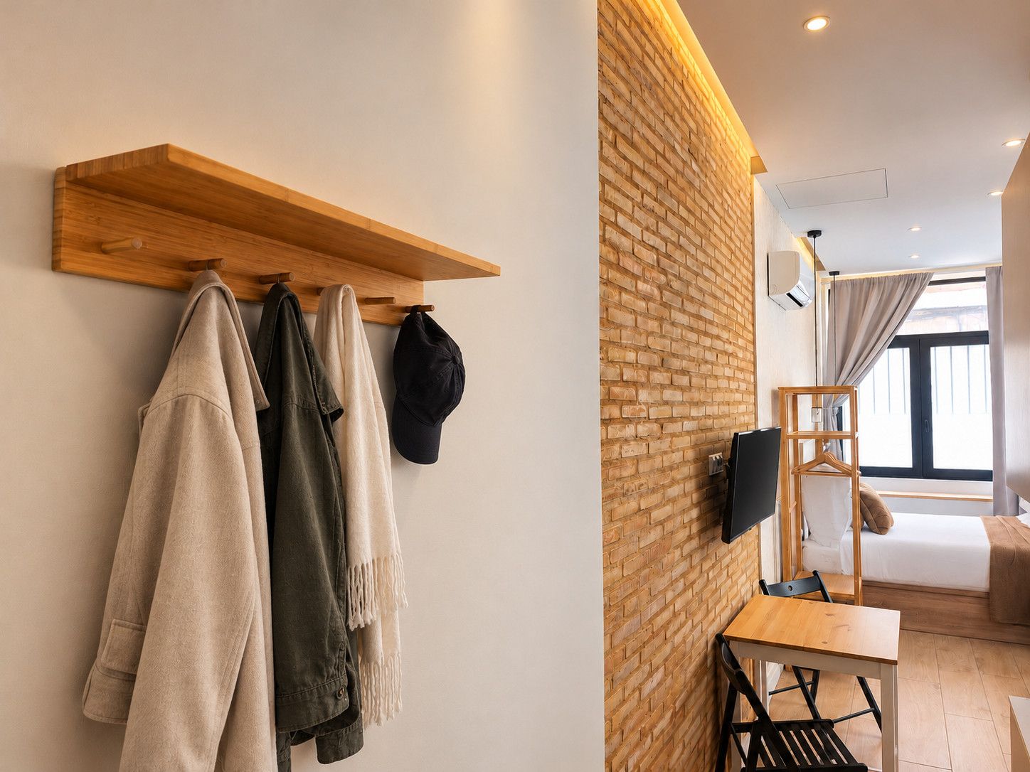 Een moderne kamer met een bakstenen muur, een houten plank met kleding, een aan de muur bevestigde televisie, een bed en een raam met gordijnen.