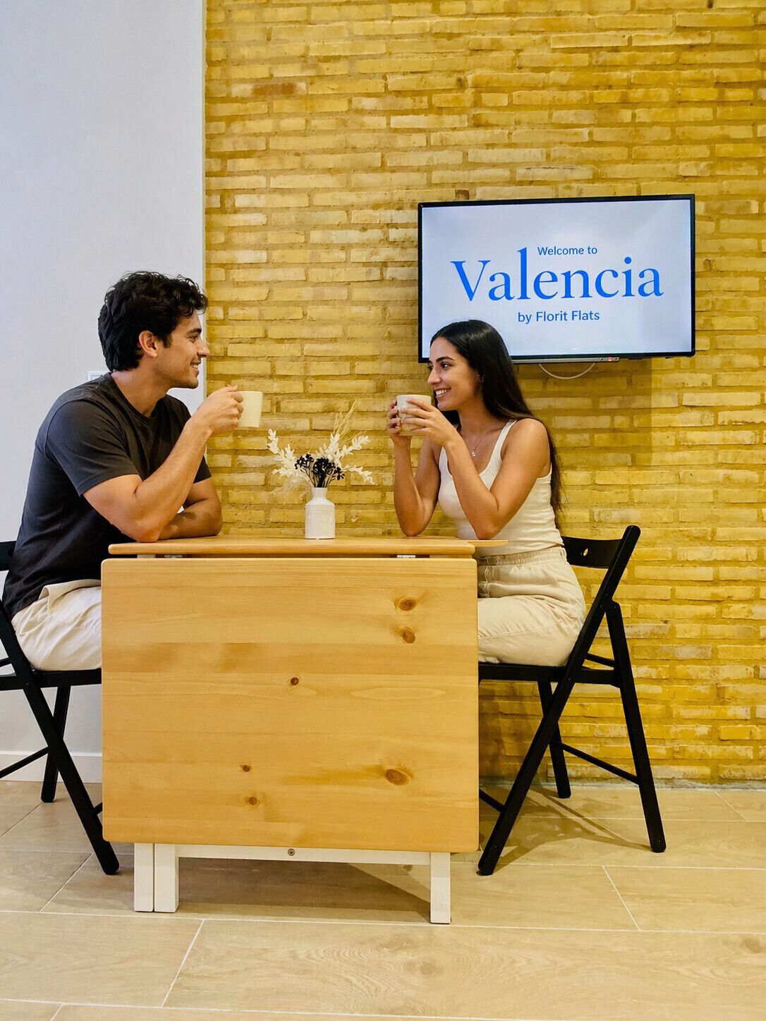 Dos personas sentadas en una mesa de madera con un jarrón de flores, frente a un muro de ladrillo con un cartel que dice 'Welcome to Valencia by Florit Flats'.