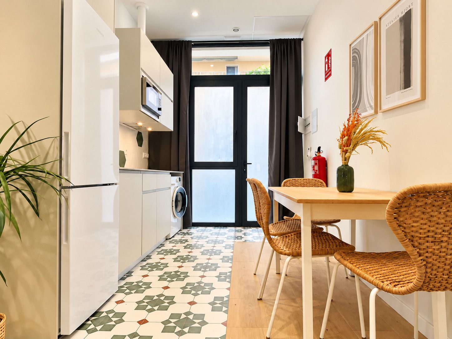 Een kleine keuken met een wasmachine, een eettafel met rieten stoelen en een vloer met een patroon.