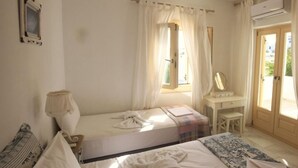 A bedroom with a bed, a desk, a chair, a lamp, and a window with curtains. - THEROS CASA LOGARAS (Paros)
