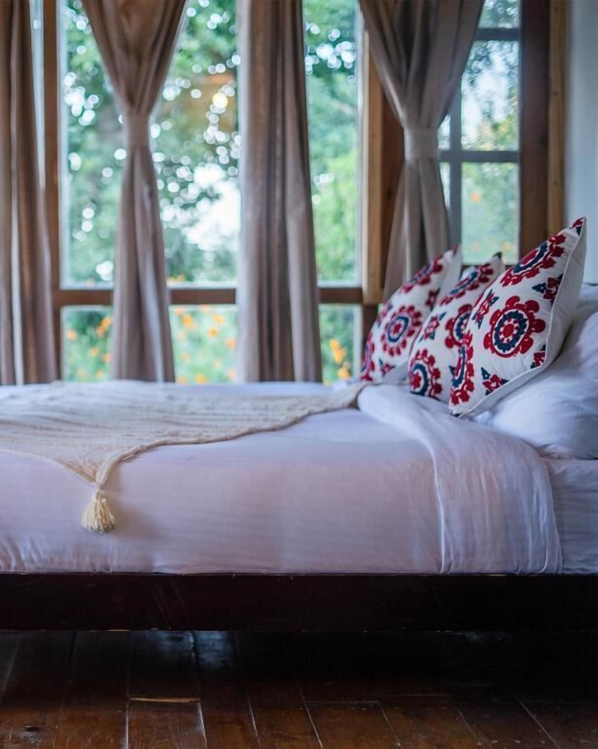 Ein Bett mit weißer Bettwäsche, gemusterten Kissen, einem Holz-Kopfteil und Blick auf Grün hinter großen, mit Vorhängen versehenen Fenstern.