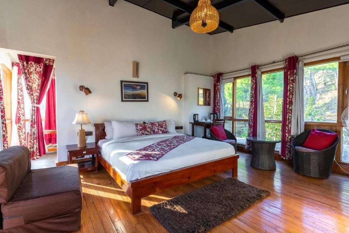 A bedroom with a wooden bed, a nightstand, a lamp, a chair, and a view of greenery through large windows.