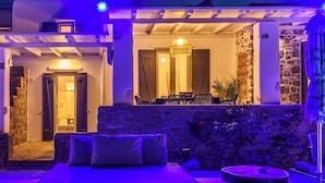 A pool area with a lounge chair and a small table, a stone wall, and a building with a door and windows in the background. - Aurora Mykonos Villas 2 (Mykonos)