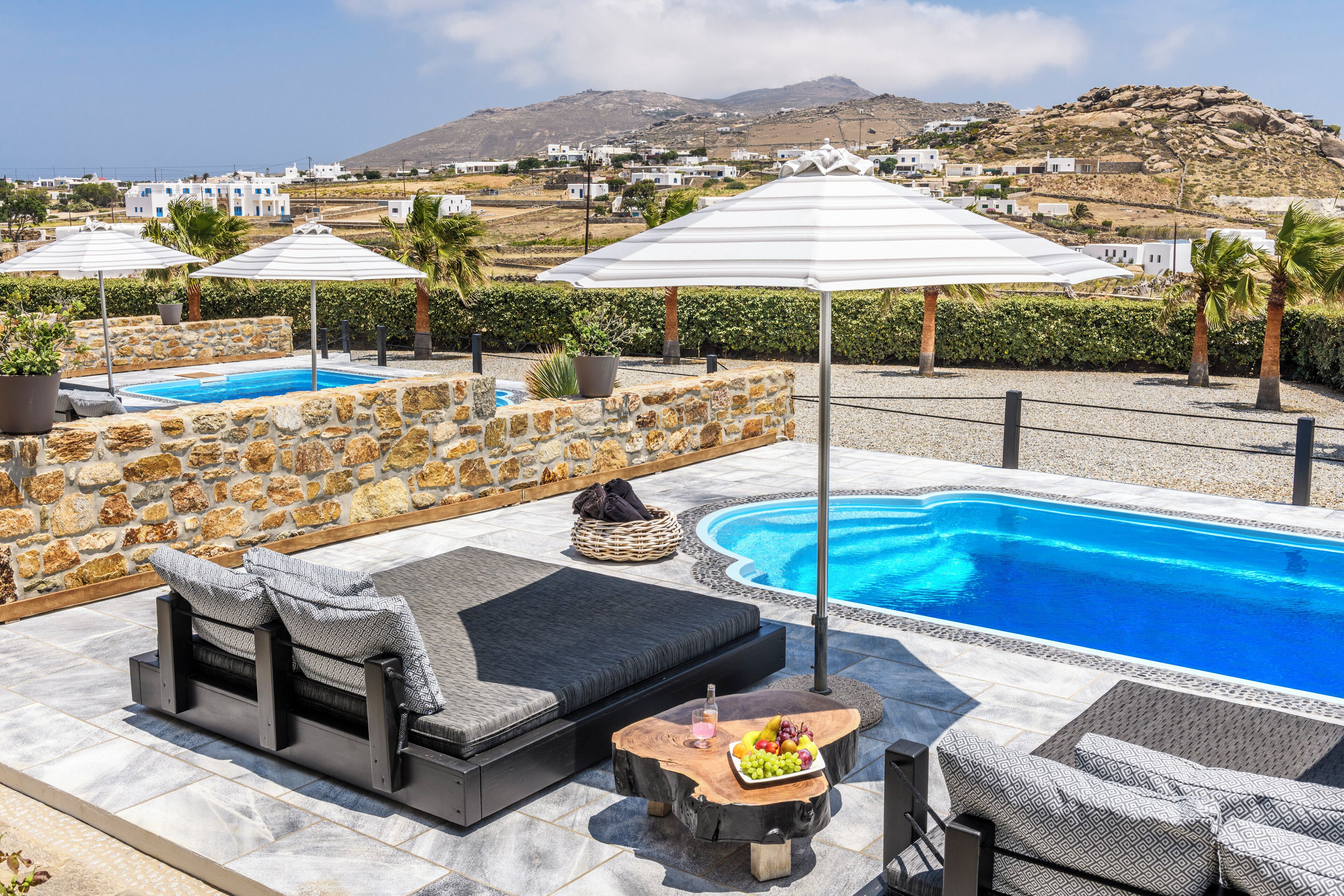 Zona de piscina con pared de piedra, sillones de descanso, sombrillas y vistas a un pueblo costero.