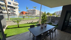 Un balcón con mesa y sillas de madera, un pequeño jardín con césped artificial y vistas a una piscina y edificios al fondo.