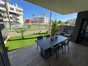 Ein Balkon mit Holztisch und -stühlen, eine kleine Gartenanlage mit Kunstrasen sowie ein Blick auf einen Pool und Gebäude im Hintergrund.