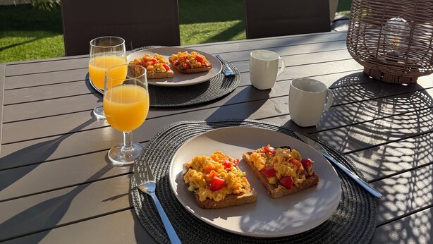 Una mesa lista para el desayuno, con tostadas, huevos revueltos y zumo de naranja, con vistas a edificios y una valla al fondo.