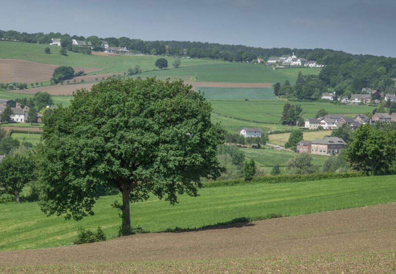 Et landligt landskab med et markant træ i forgrunden, bølgende marker og spredte huse i baggrunden.