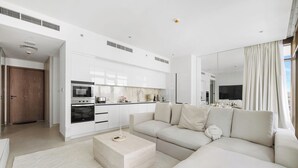 A modern living room with a beige sofa, a coffee table, and a kitchen area with white cabinets and appliances. - Serene 1BR Escape with Burj Khalifa View (Dubai)